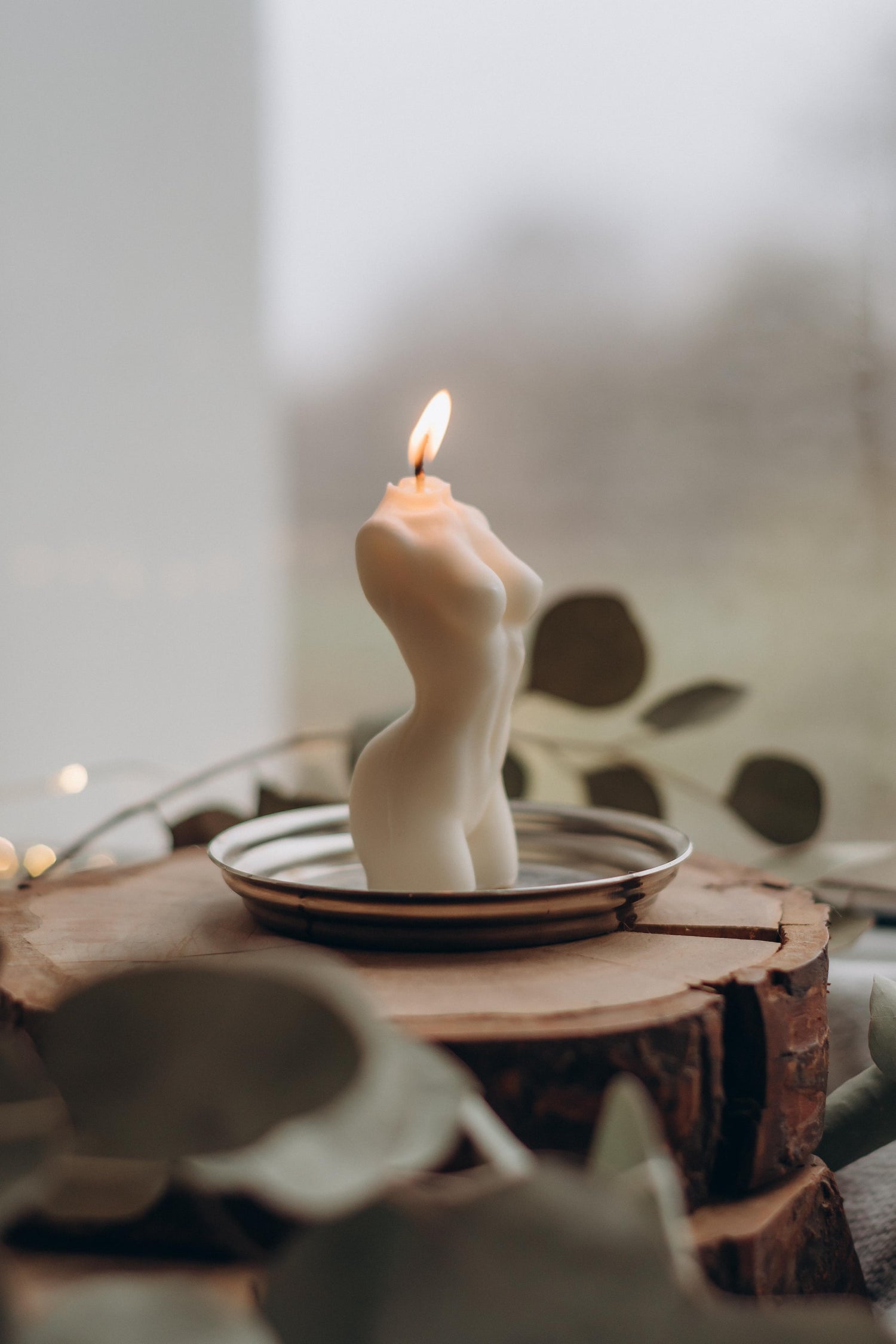 CandlesCo lit candle of nude woman on a tin plate sitting on a timber base surrounded by focal blurred leaves  